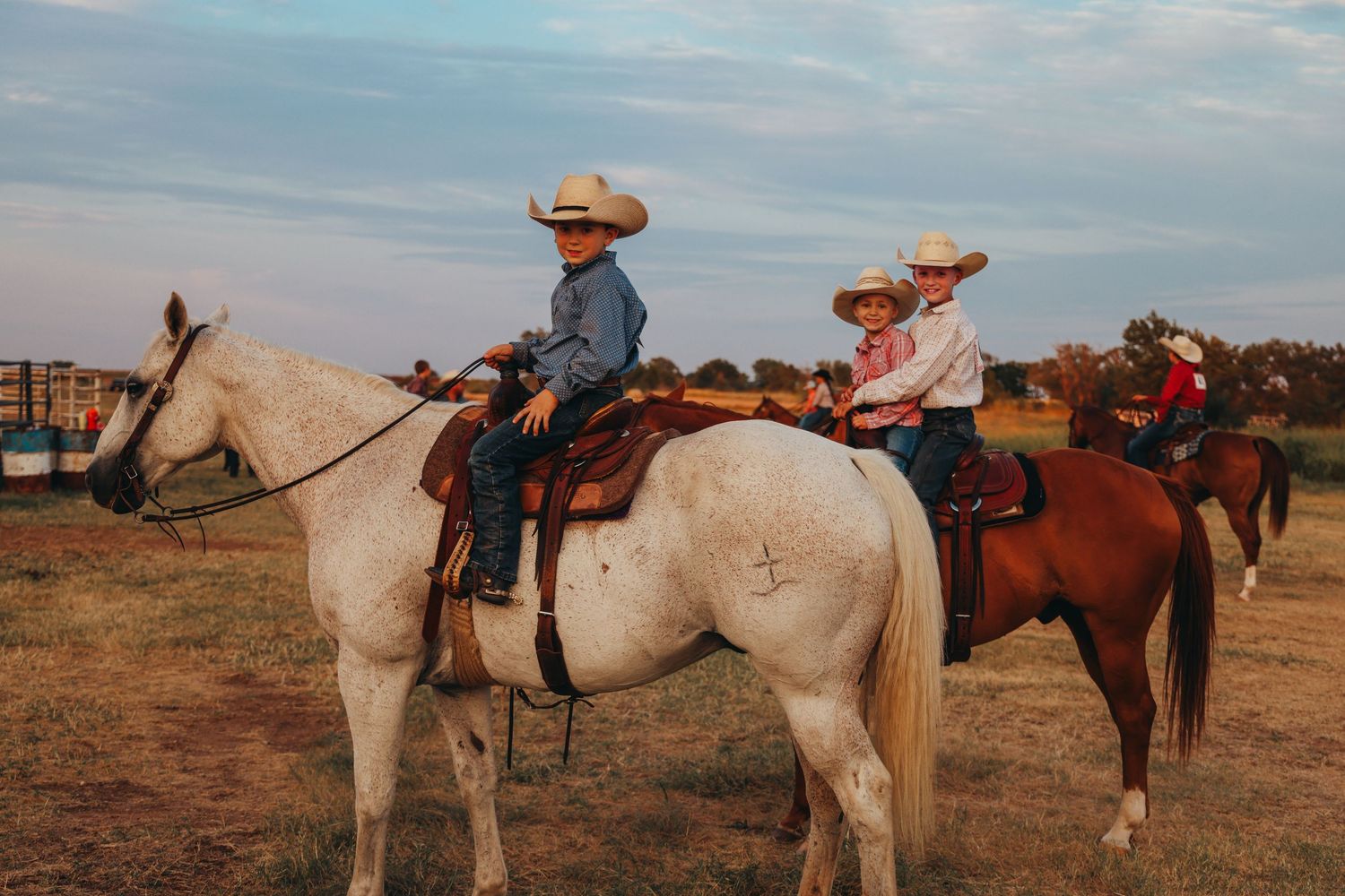 White and brown horses with riders wearing cowboy hats against sunset sky.