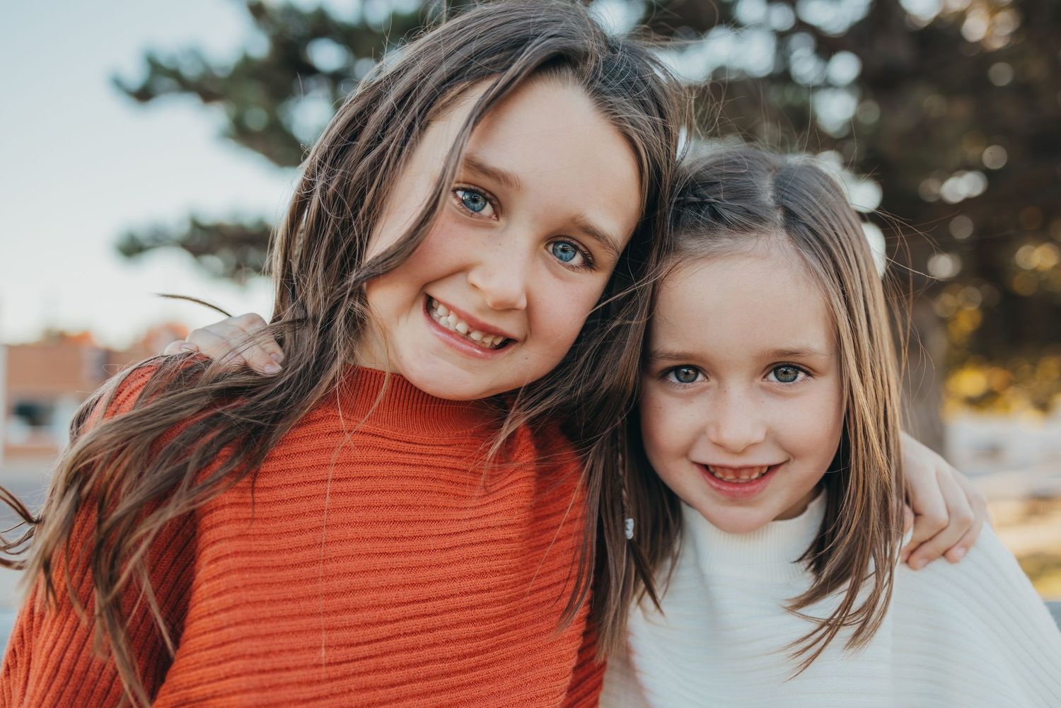 Two smiling girls with brown hair pose together outdoors, wearing red and white sweaters in a warm family portrait.