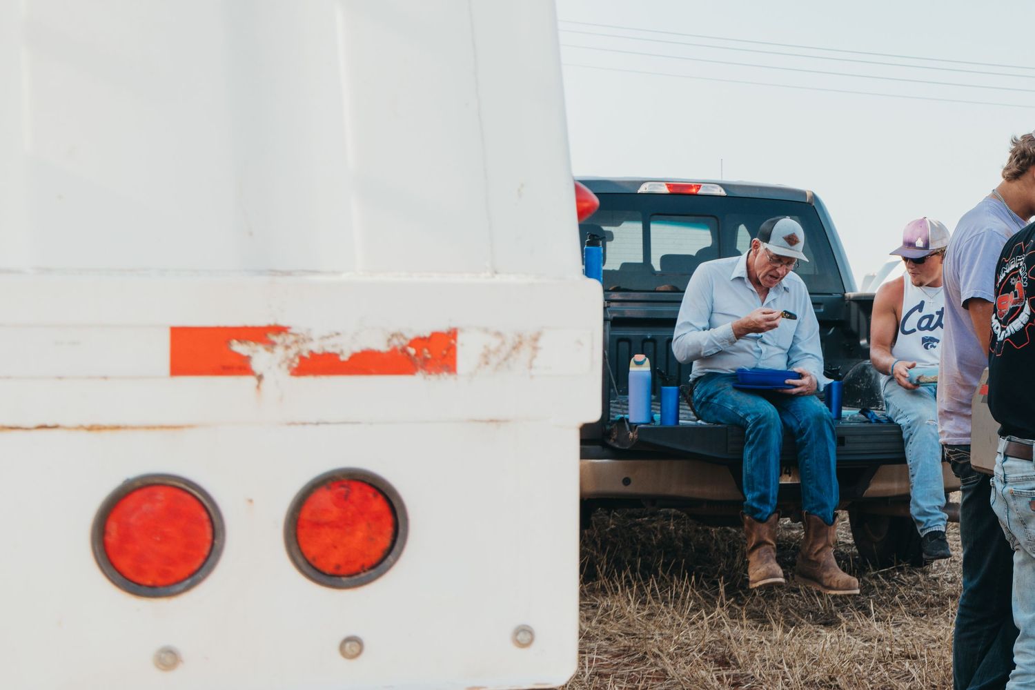 People gathered near a white vehicle with red tail lights in a rural field setting.