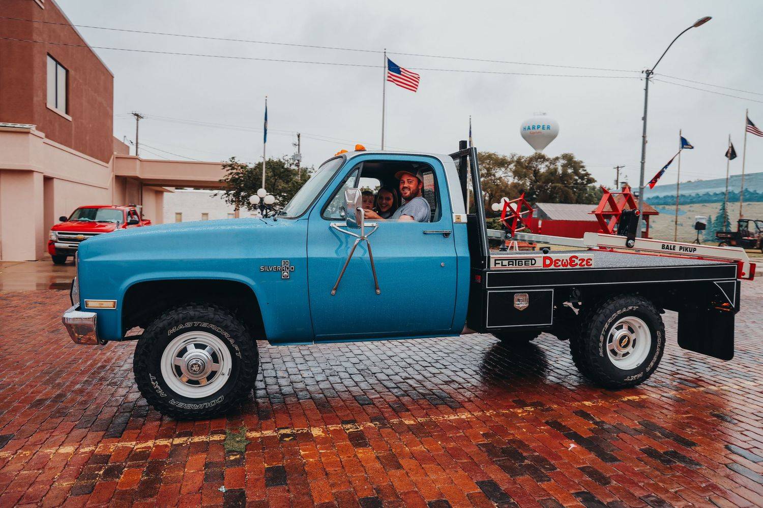 Classic blue Chevrolet flatbed truck parked on wet brick street with American flag.