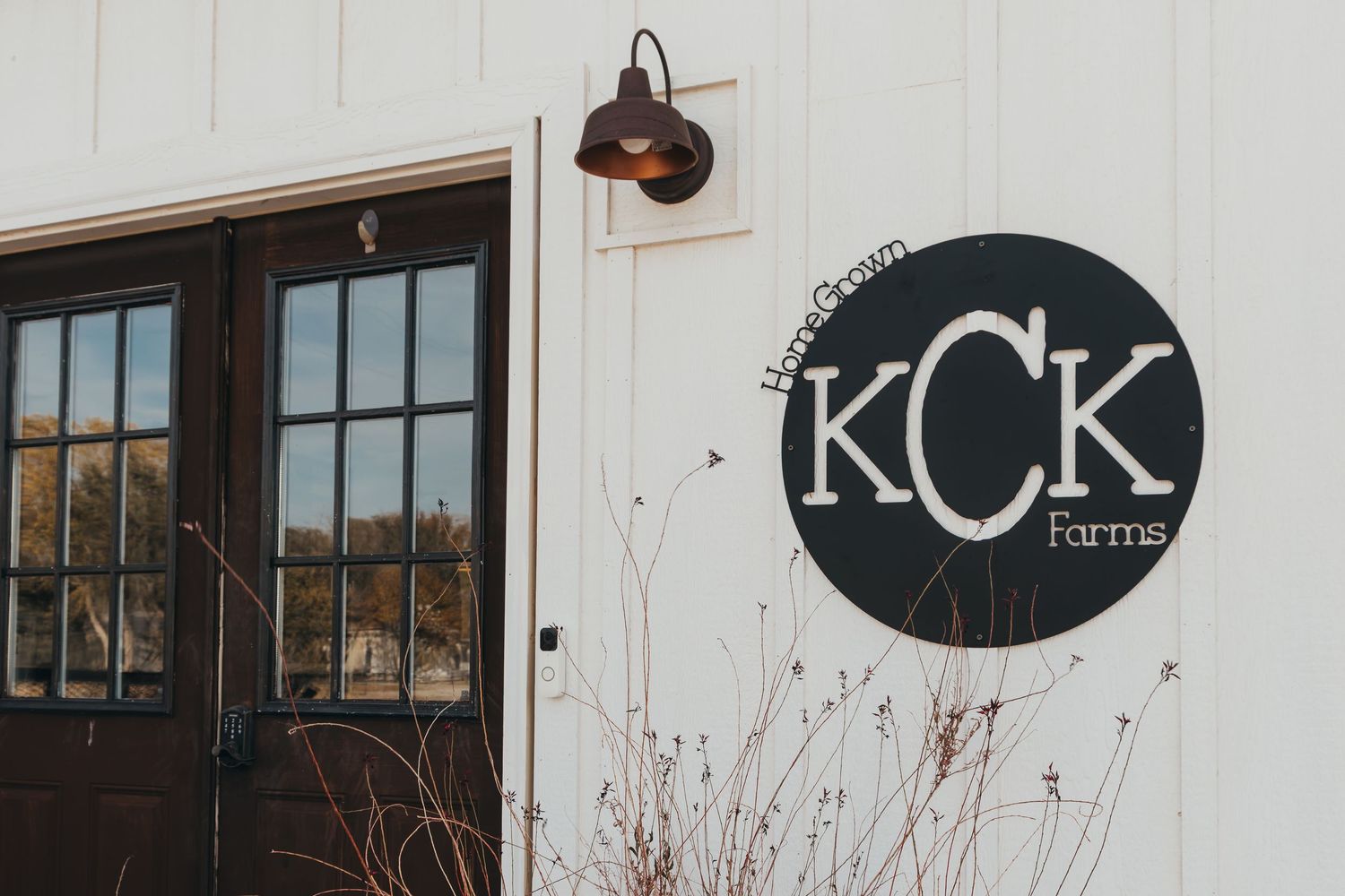 Black and white KCK Farms circular logo sign mounted on white wall next to dark-framed glass door with rustic sconce light.