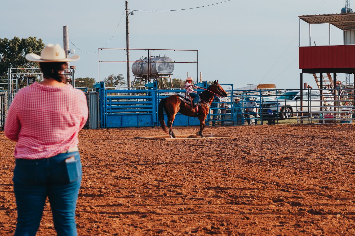 Cowboy competes in rodeo event at dusty arena while spectator in pink shirt watches from sidelines.