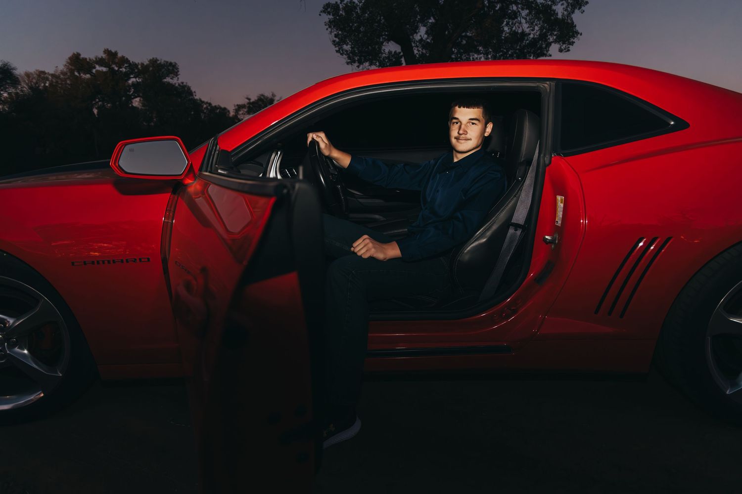 A red sports car with an open door at dusk, with someone sitting in the driver's seat wearing dark clothing.