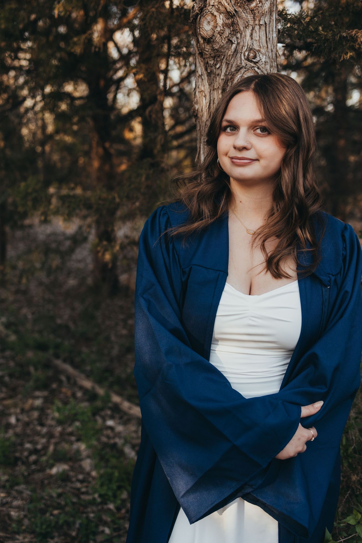 Young woman in white dress and blue graduation gown leaning against tree in forest setting.