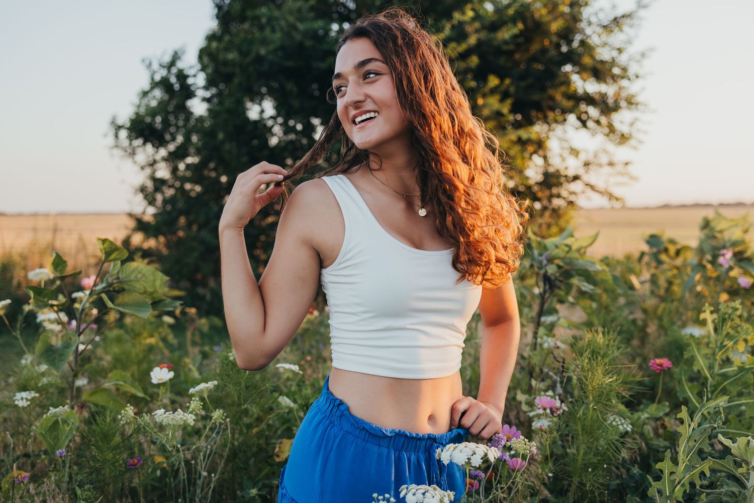 A smiling woman in a white crop top and blue skirt stands in a wildflower field during golden hour.