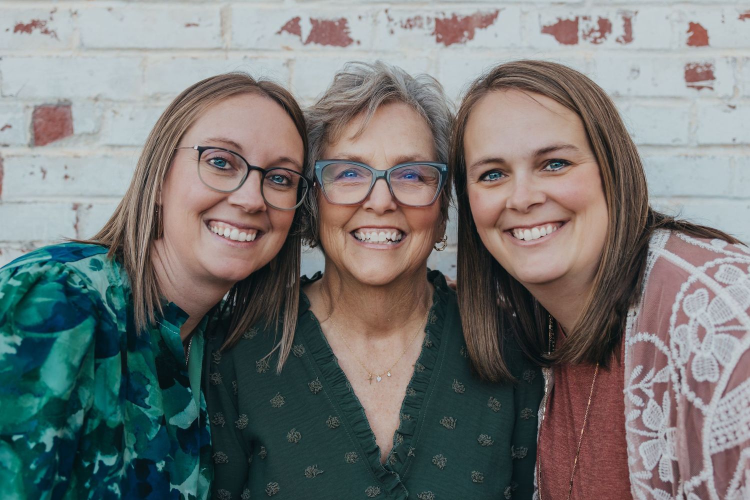Three smiling women pose together in front of a white brick wall in a warm, joyful family portrait.