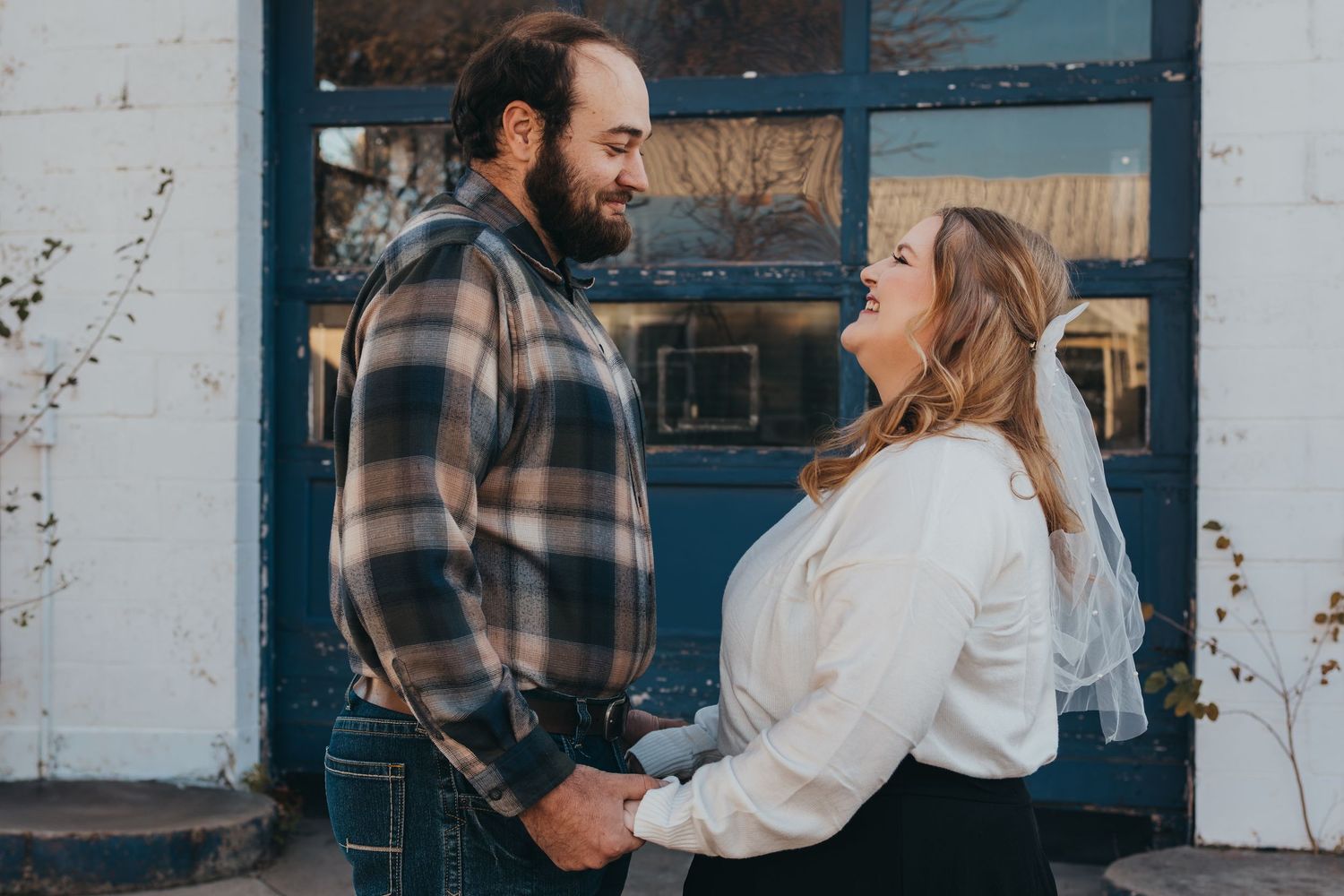 A couple in casual winter attire shares an intimate moment against a rustic white wall and blue door background.