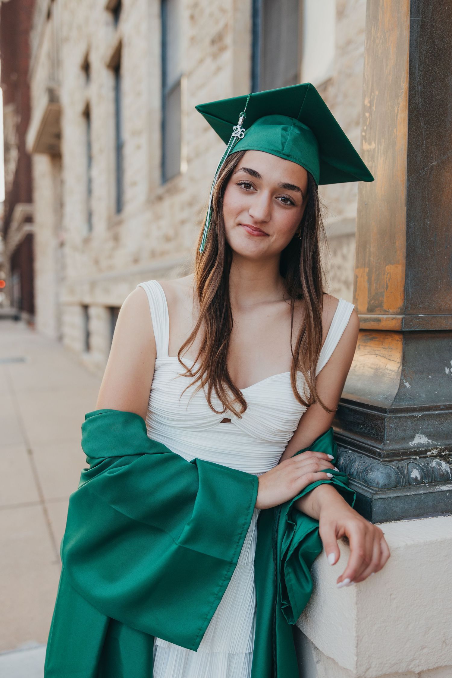 Young woman in green graduation cap and gown smiling outdoors near a stone column in a white dress.