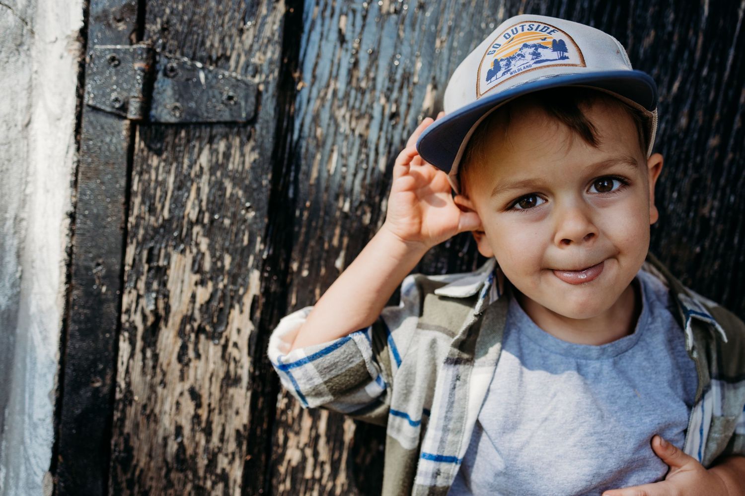 Young child in a baseball cap and denim jacket smiles brightly while standing against weathered wooden door.
