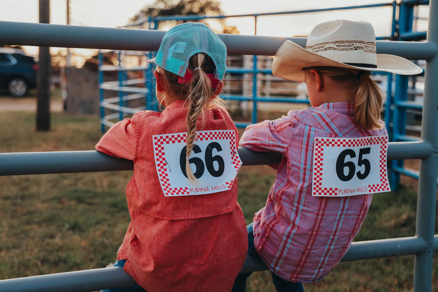 Two competitors wearing numbered bibs 65 and 66 lean against rodeo arena fence in western attire.