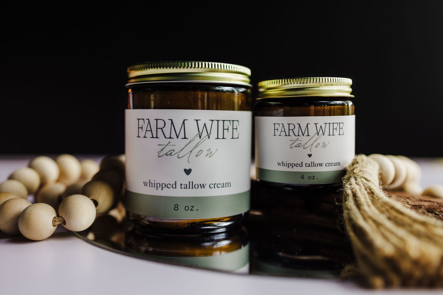 Two glass jars of Farm Wife branded whipped tallow cream products displayed with wooden beads and twine on white surface.