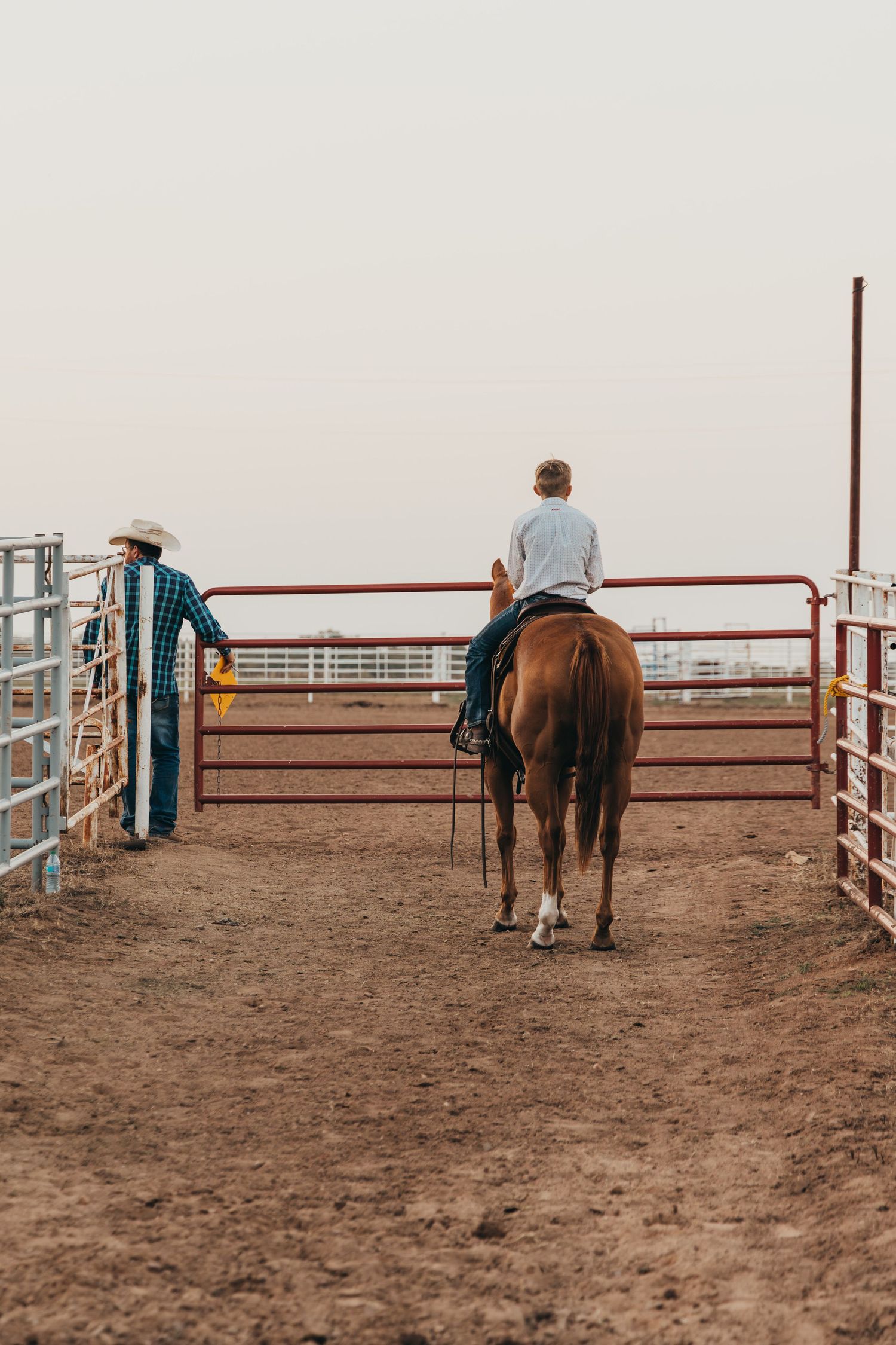 Two horses and riders stand in a dusty arena with metal corral fencing under an overcast sky.