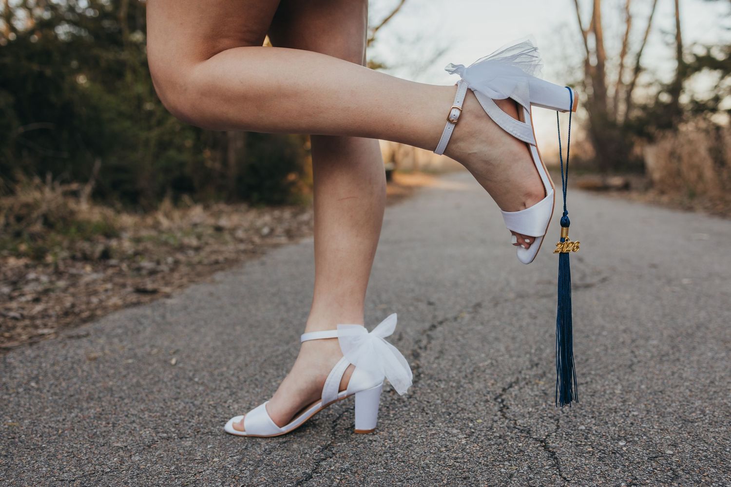 Close-up of white block heel sandals with bow detail and graduation tassel accessory on a road.