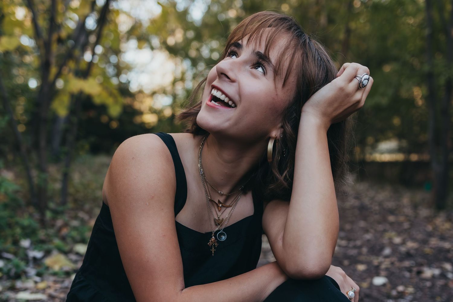 A person in a black tank top laughs joyfully while sitting outdoors among autumn leaves and trees.