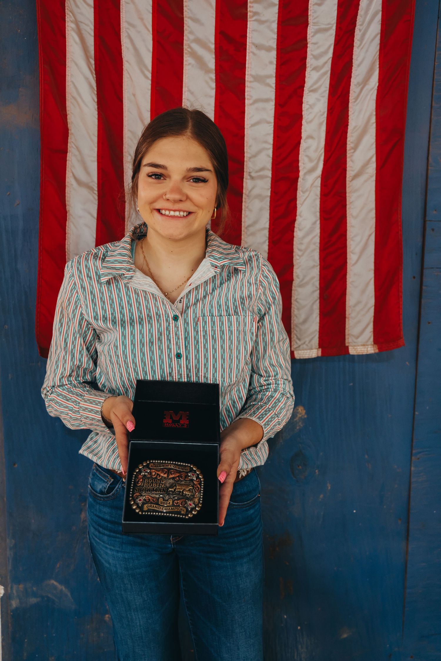 Student in striped shirt and jeans stands proudly holding award certificate in front of American flag backdrop.