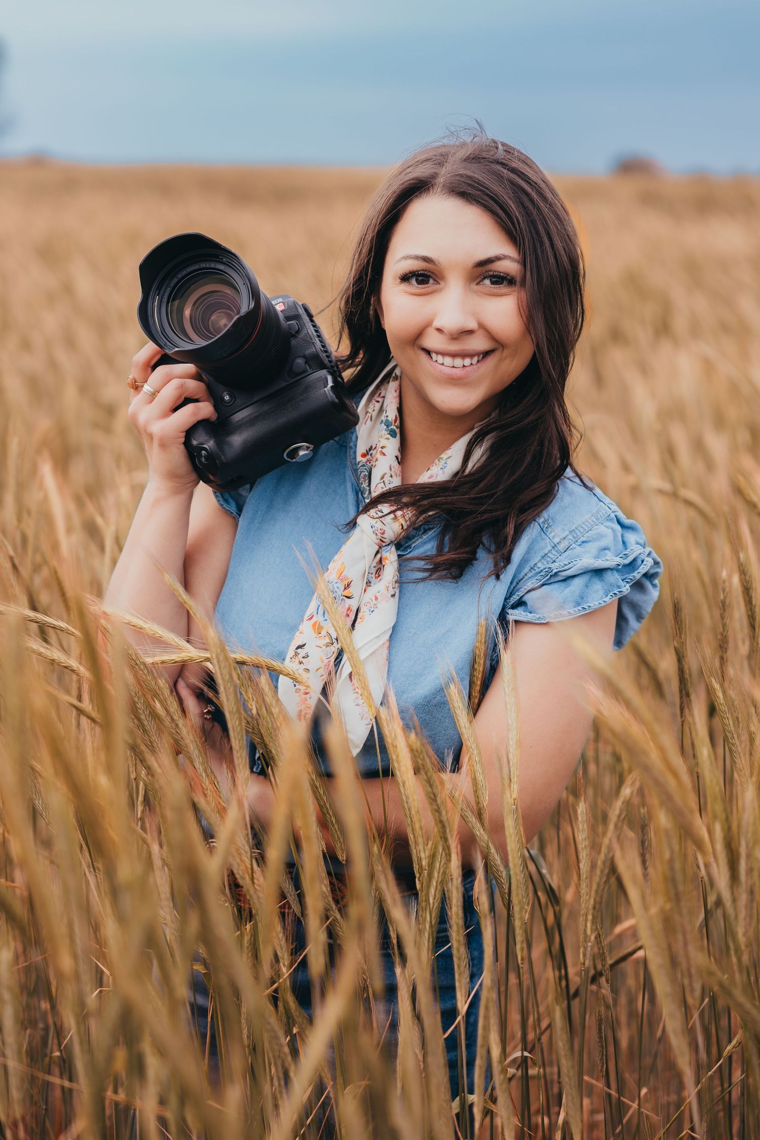 A photographer holds a DSLR camera while standing in a golden wheat field wearing a blue top and printed scarf.