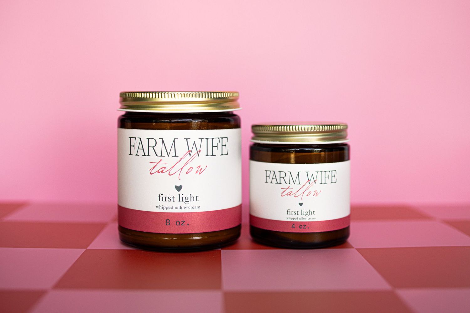Two Farm Wife Tallow 'First Light' amber glass jars with gold lids displayed on a pink checkered surface.