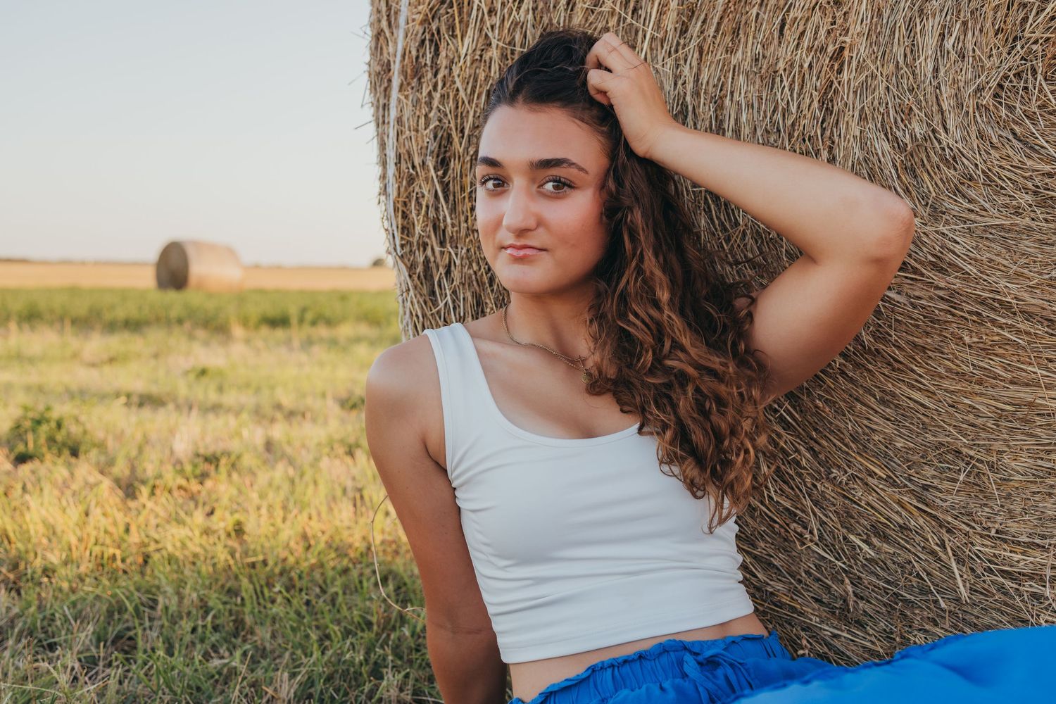 Someone leans against a golden hay bale in a rural field during sunset, wearing a white tank top and blue bottoms.