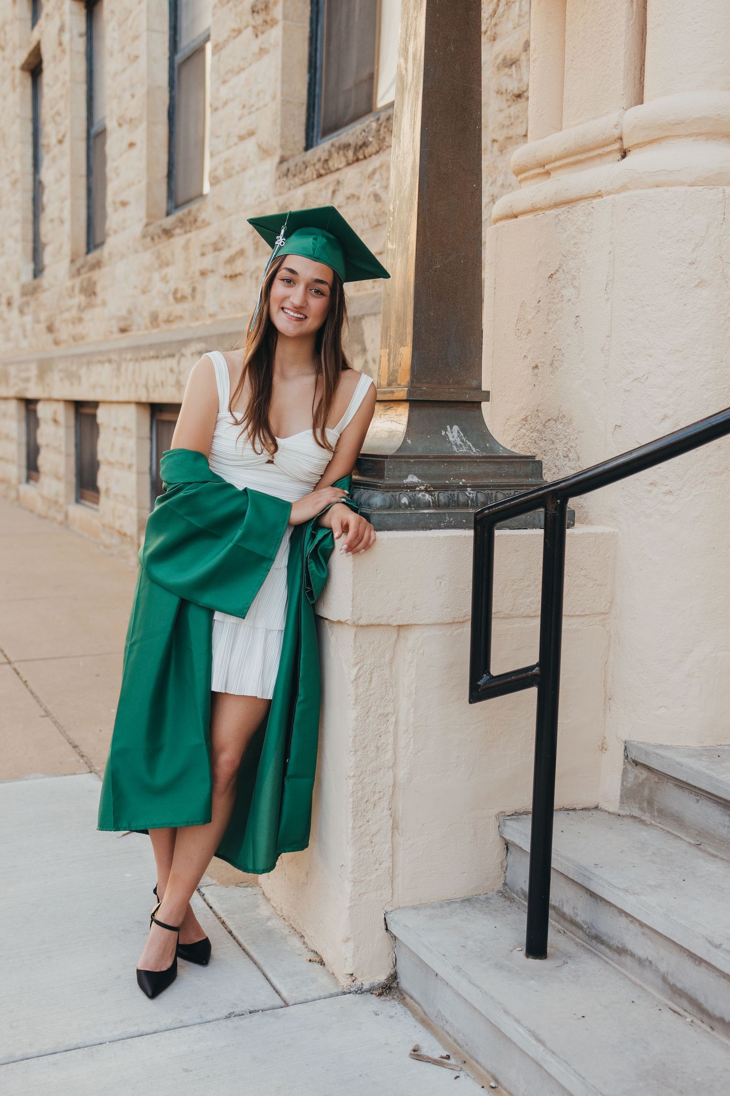Young woman in green graduation cap and gown poses proudly on steps outside a stone building.