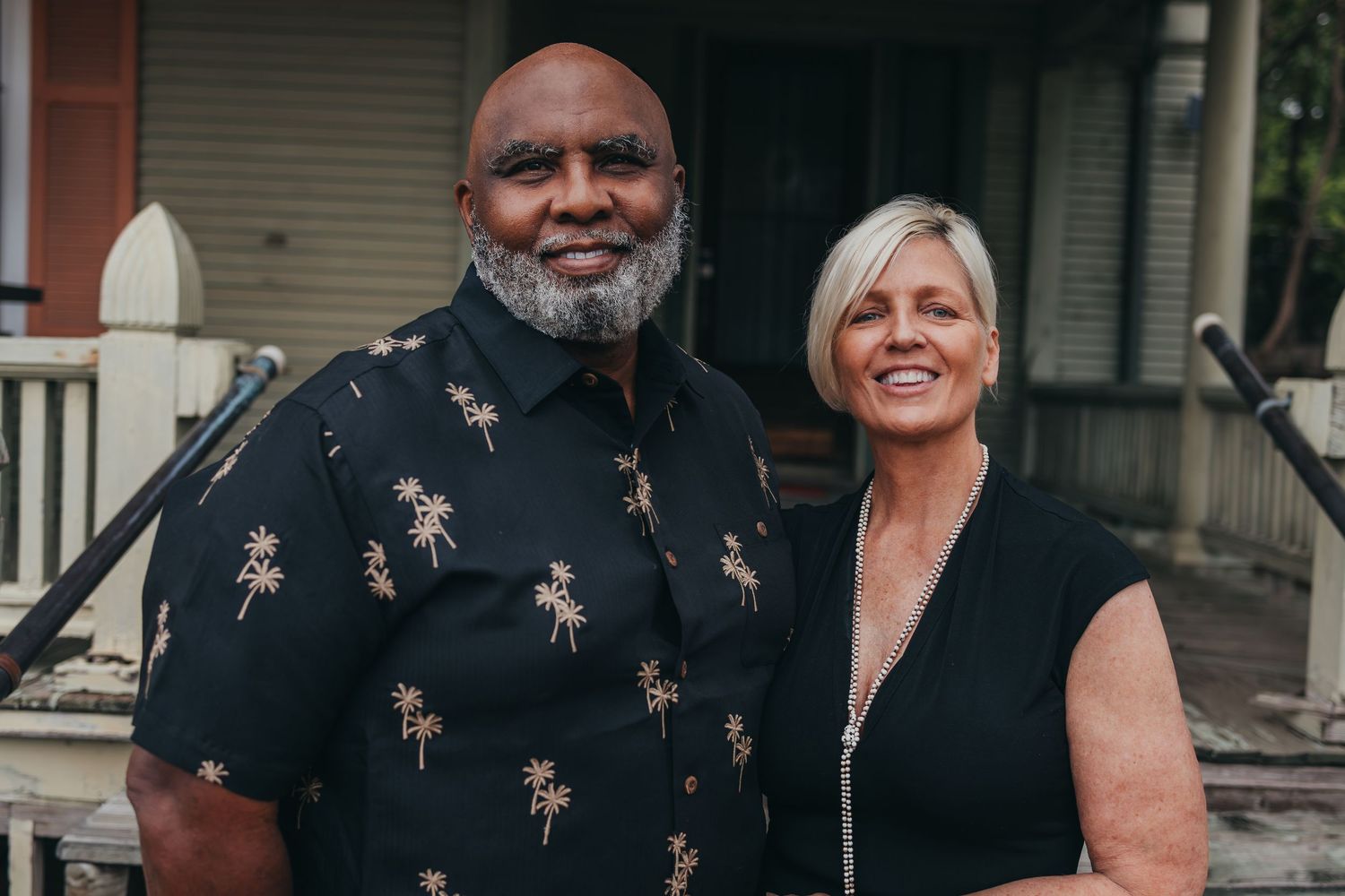 A beaming couple stands side by side on a porch, the man wearing a dark shirt with palm tree pattern and woman in black dress.