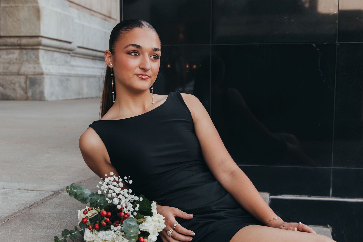 A person in a black dress poses elegantly with a bouquet of flowers against a stone wall background.