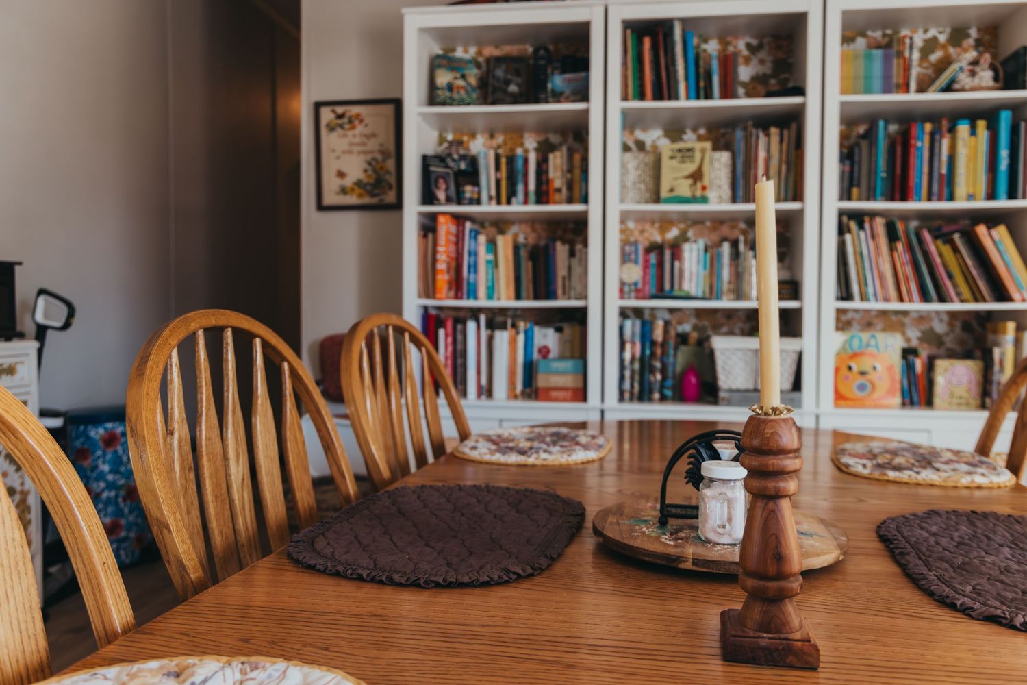 A cozy dining room setup with wooden chairs and white bookshelves filled with books and decor in the background.