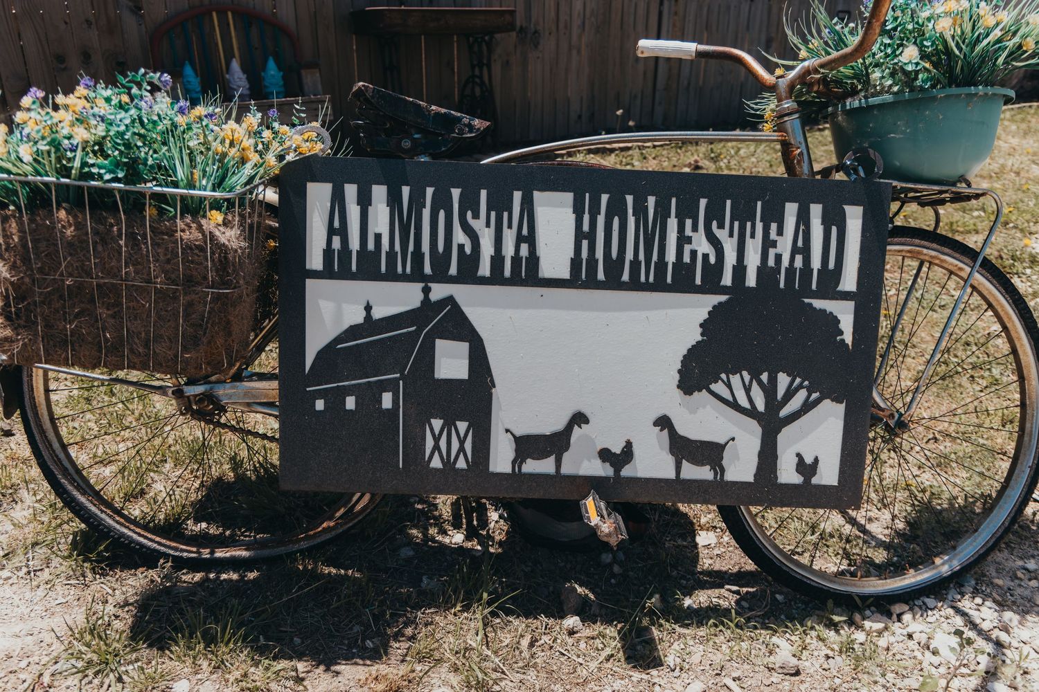 Black metal farmhouse sign with silhouettes of a barn and animals mounted on a vintage bicycle against a rustic backdrop.