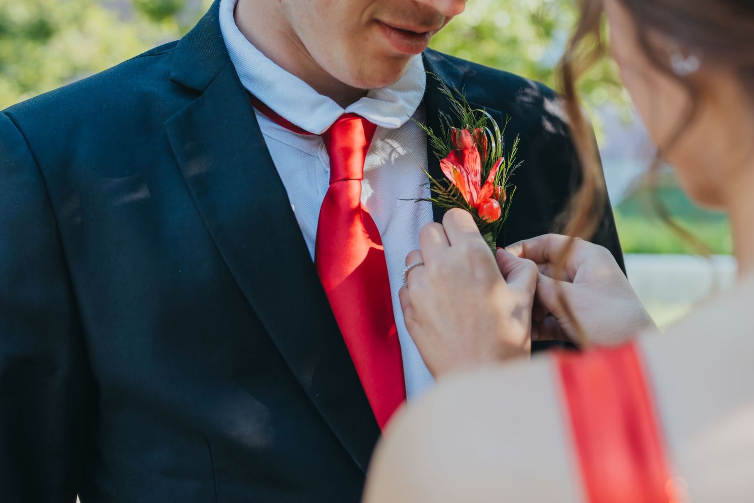 Someone adjusts a vibrant red boutonniere with tropical flowers on a navy suit jacket with matching red tie.
