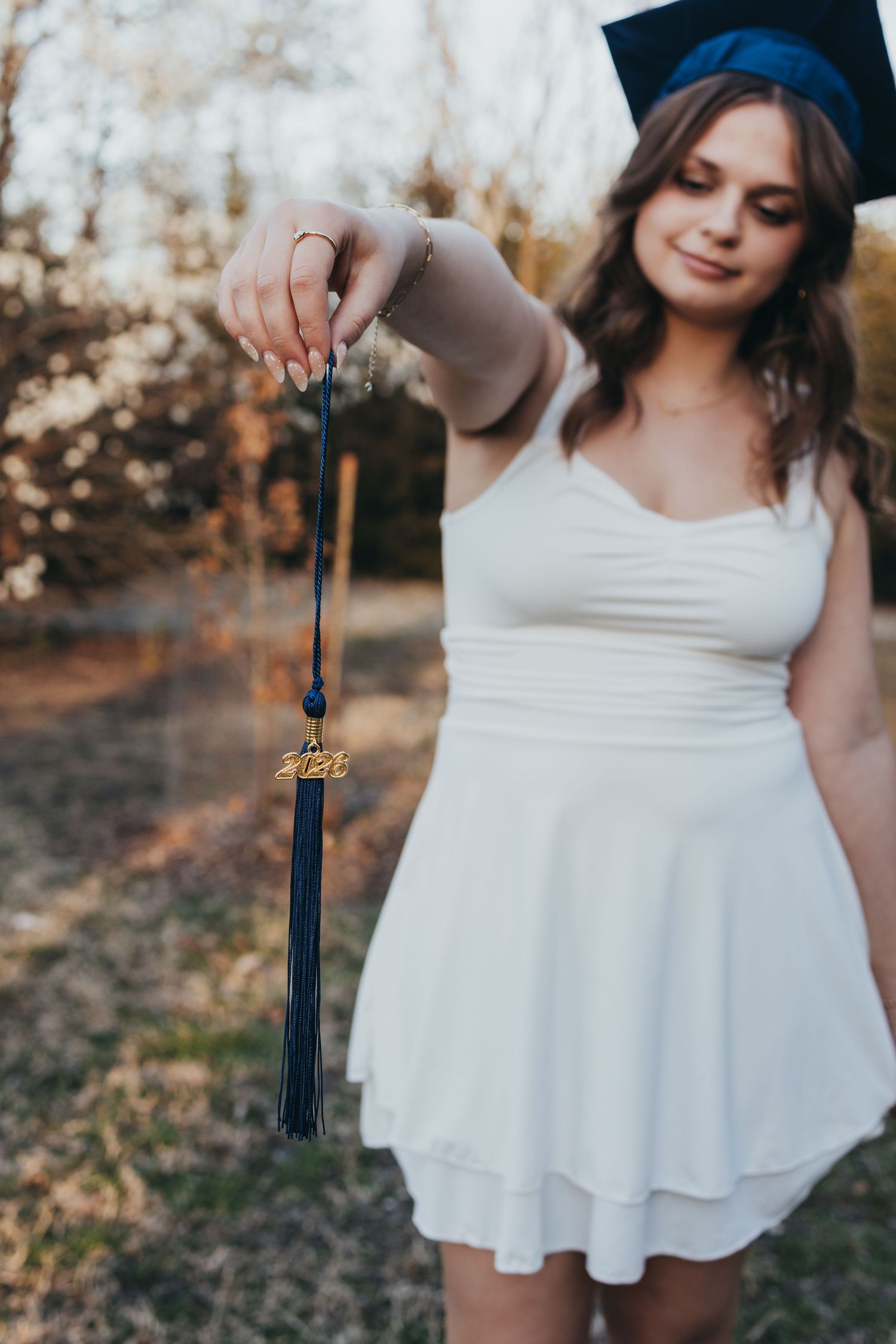 Young woman in white dress and blue graduation cap holds tassel with 2023 charm outdoors.