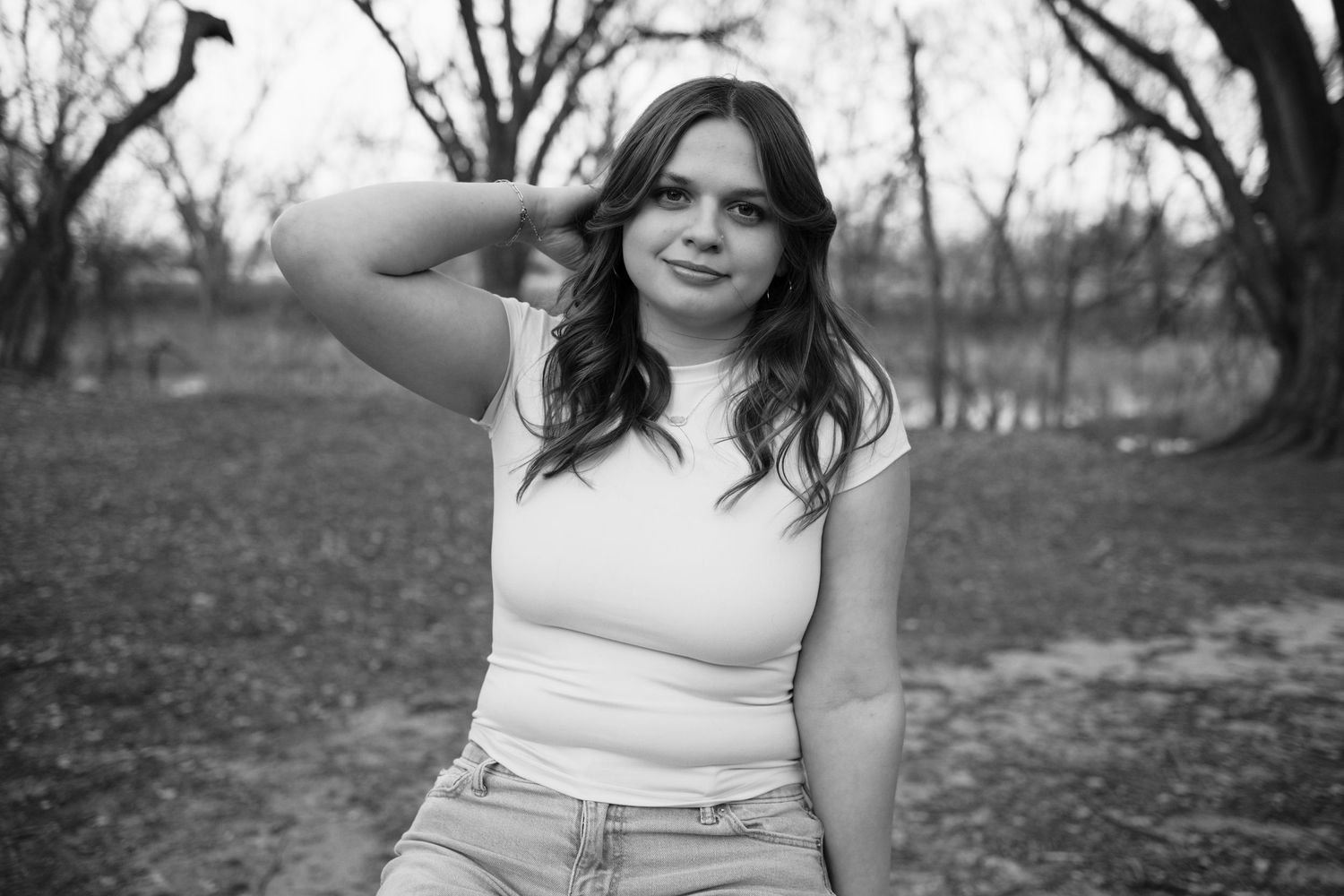 Black and white portrait of a young woman in a white top posing outdoors among bare trees.