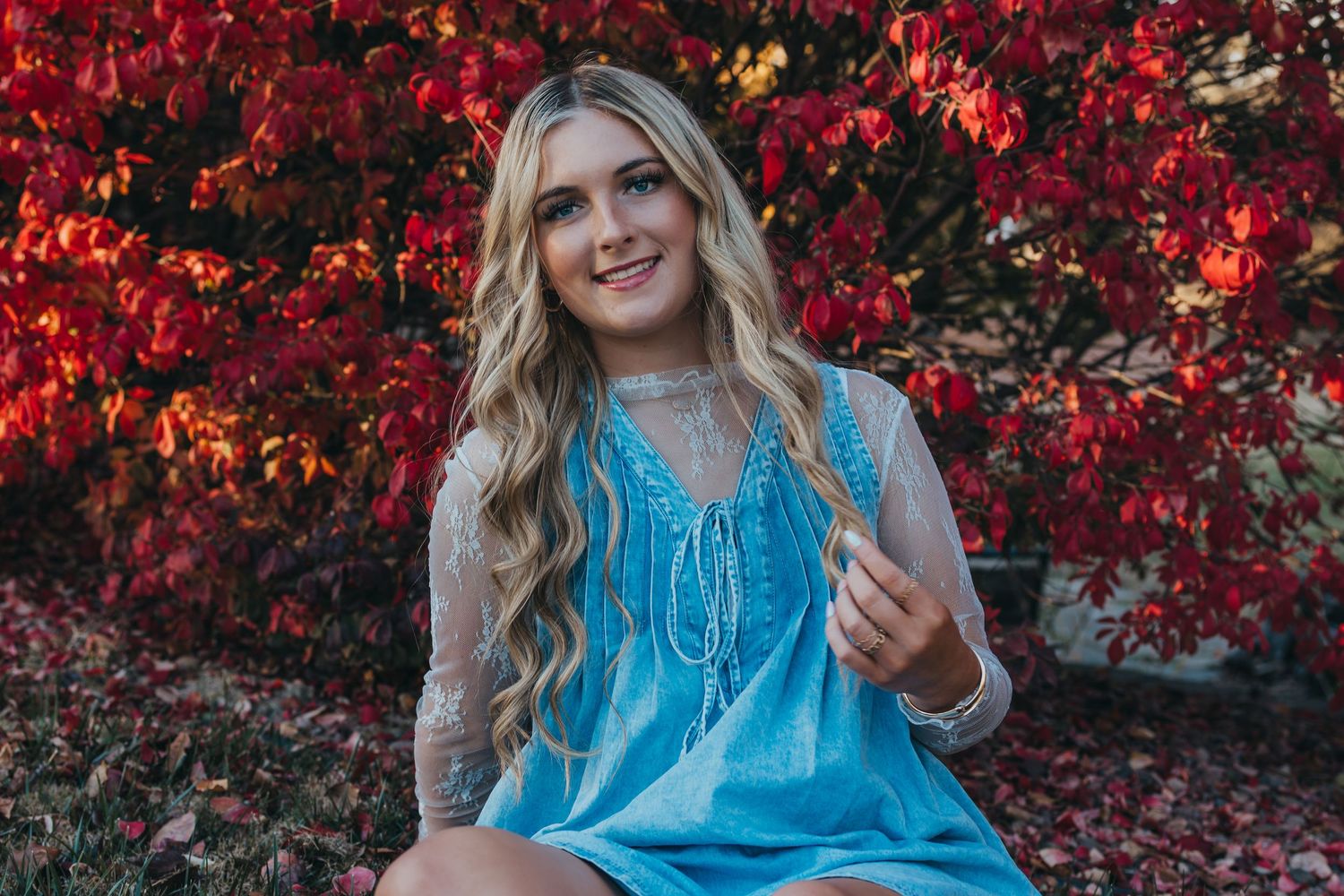 Young blonde woman in blue dress smiling outdoors against vibrant red autumn foliage background.
