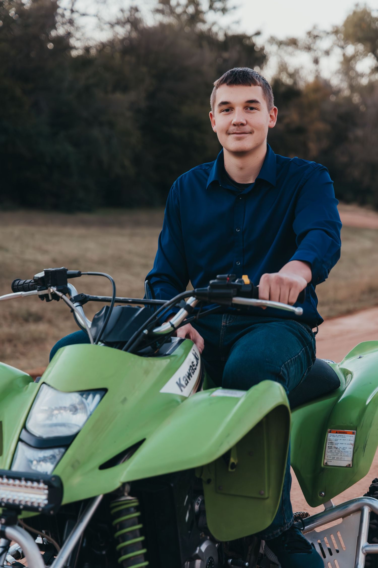 Young man in navy shirt smiling while sitting on a green Kawasaki ATV quad bike outdoors.