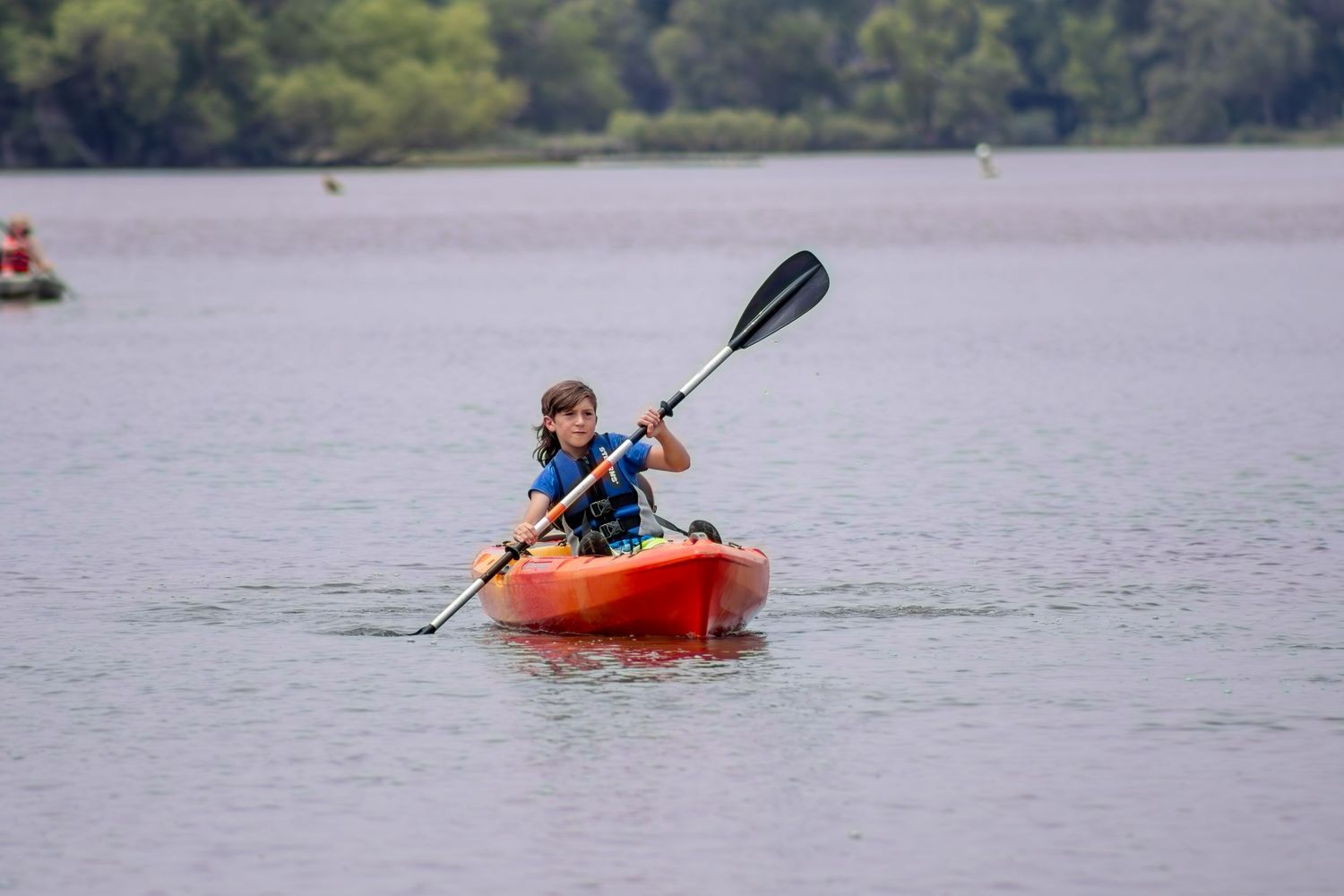 A kayaker paddles an orange kayak across tranquil waters with trees in the background.