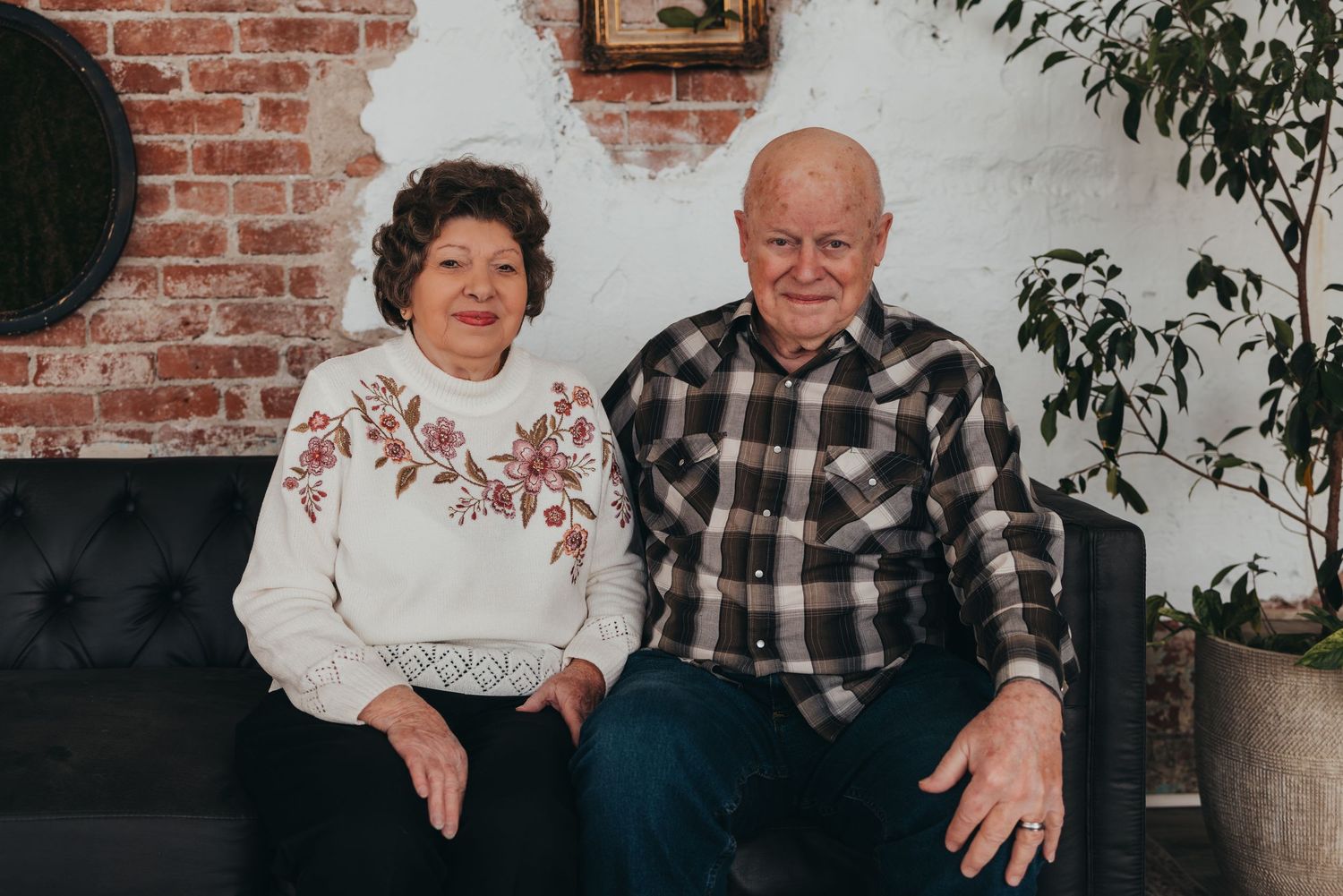 Senior couple sitting together on couch near brick wall and indoor plants.