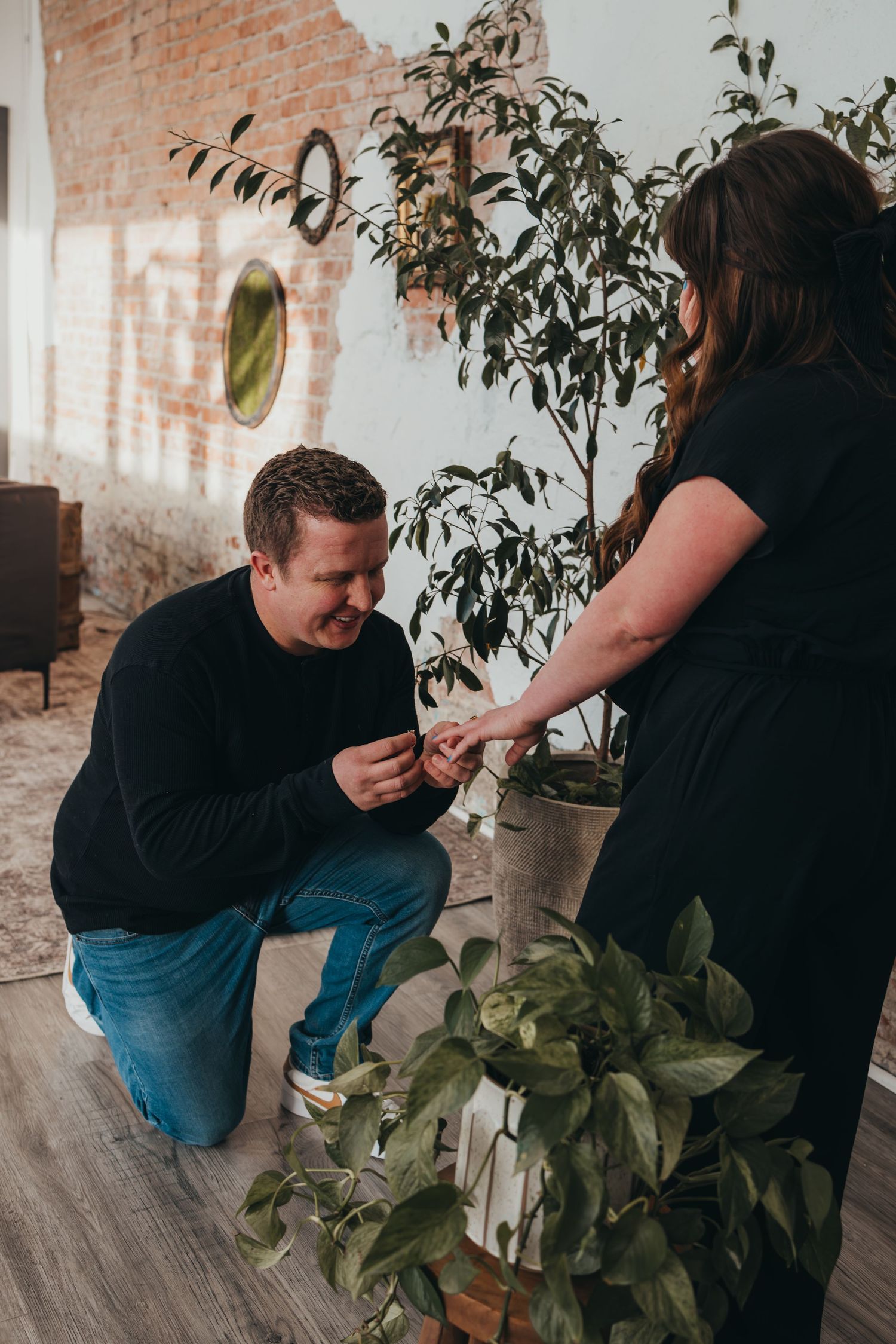 Person in black sweater kneeling to propose in room with brick wall and plants.