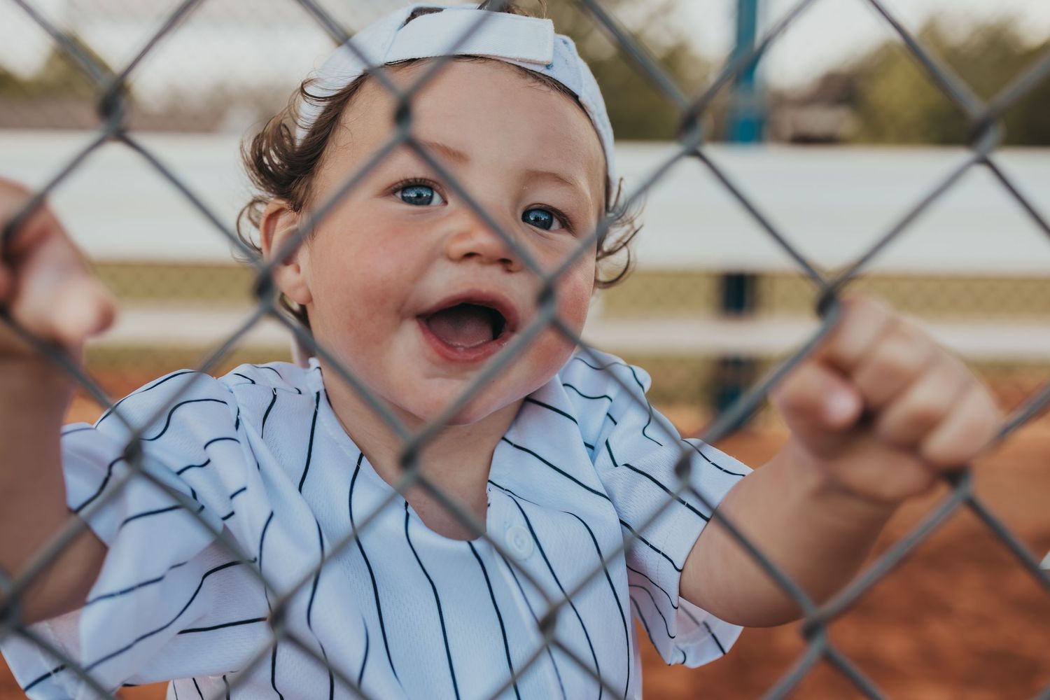 A cheerful child in baseball attire smiles while holding onto a chain-link fence.