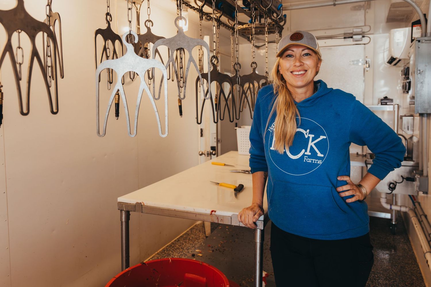 A person wearing a blue CK sweatshirt smiles while working in a kitchen prep area with utensils hanging on the wall.