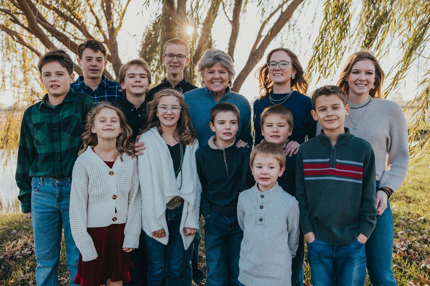 A large group of individuals in casual clothing poses together outdoors in front of autumn trees.