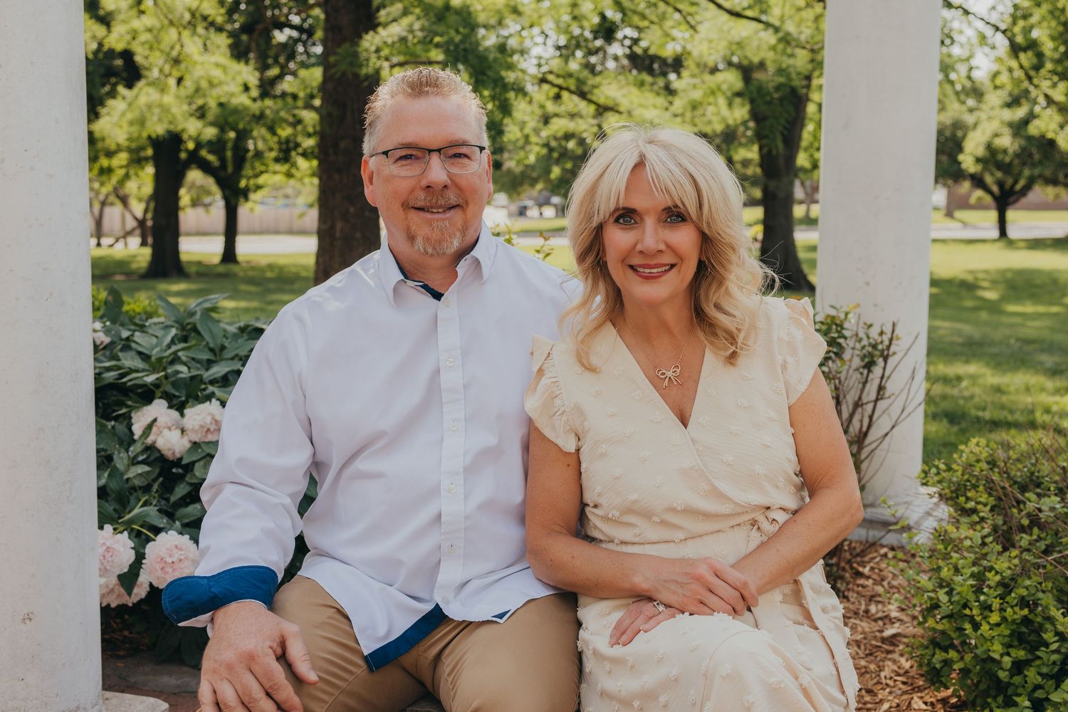A couple sits together on outdoor porch steps wearing light colored clothing in a garden setting during summer.