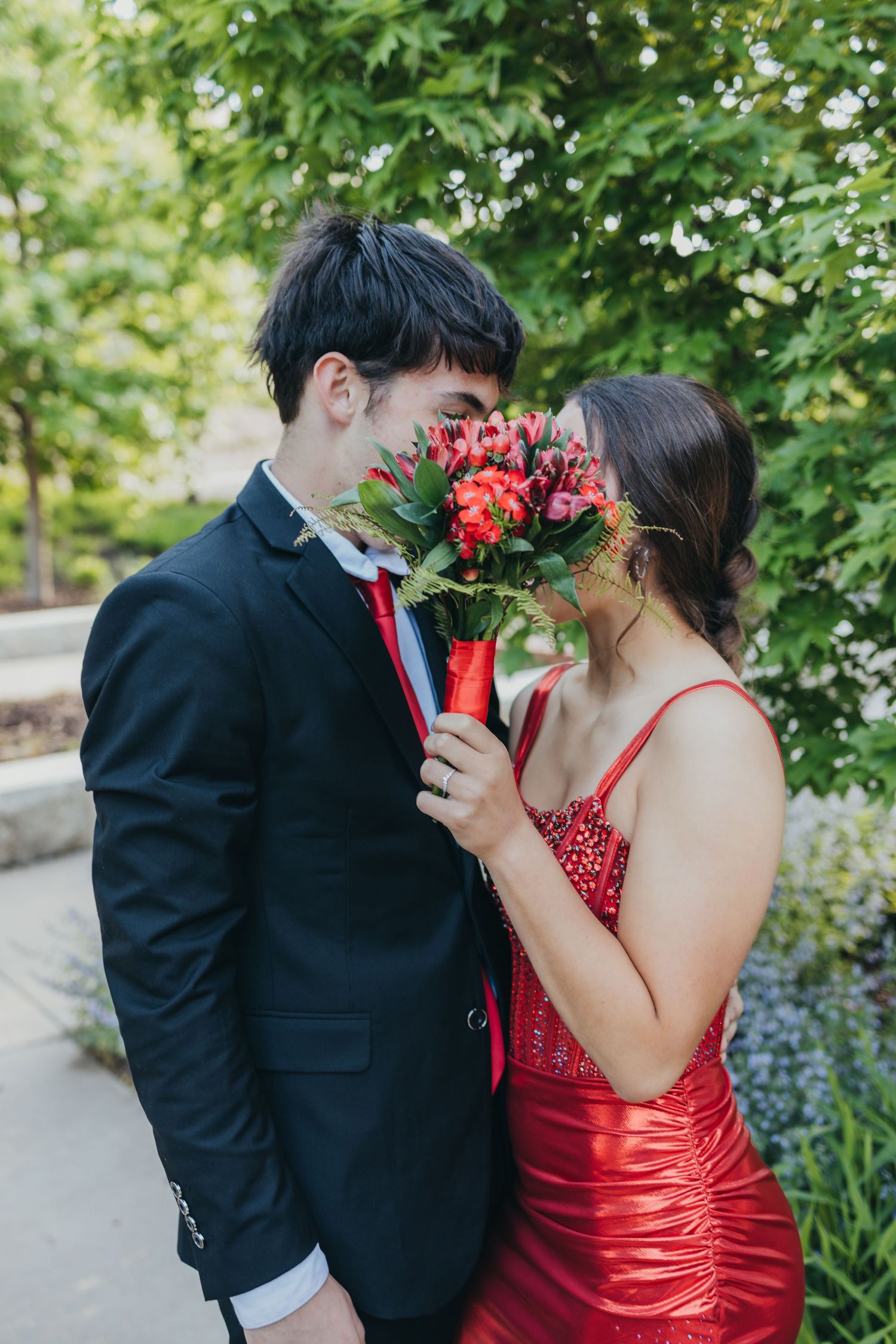 Couple sharing intimate moment with red roses in front of lush greenery, wearing formal attire for special occasion.