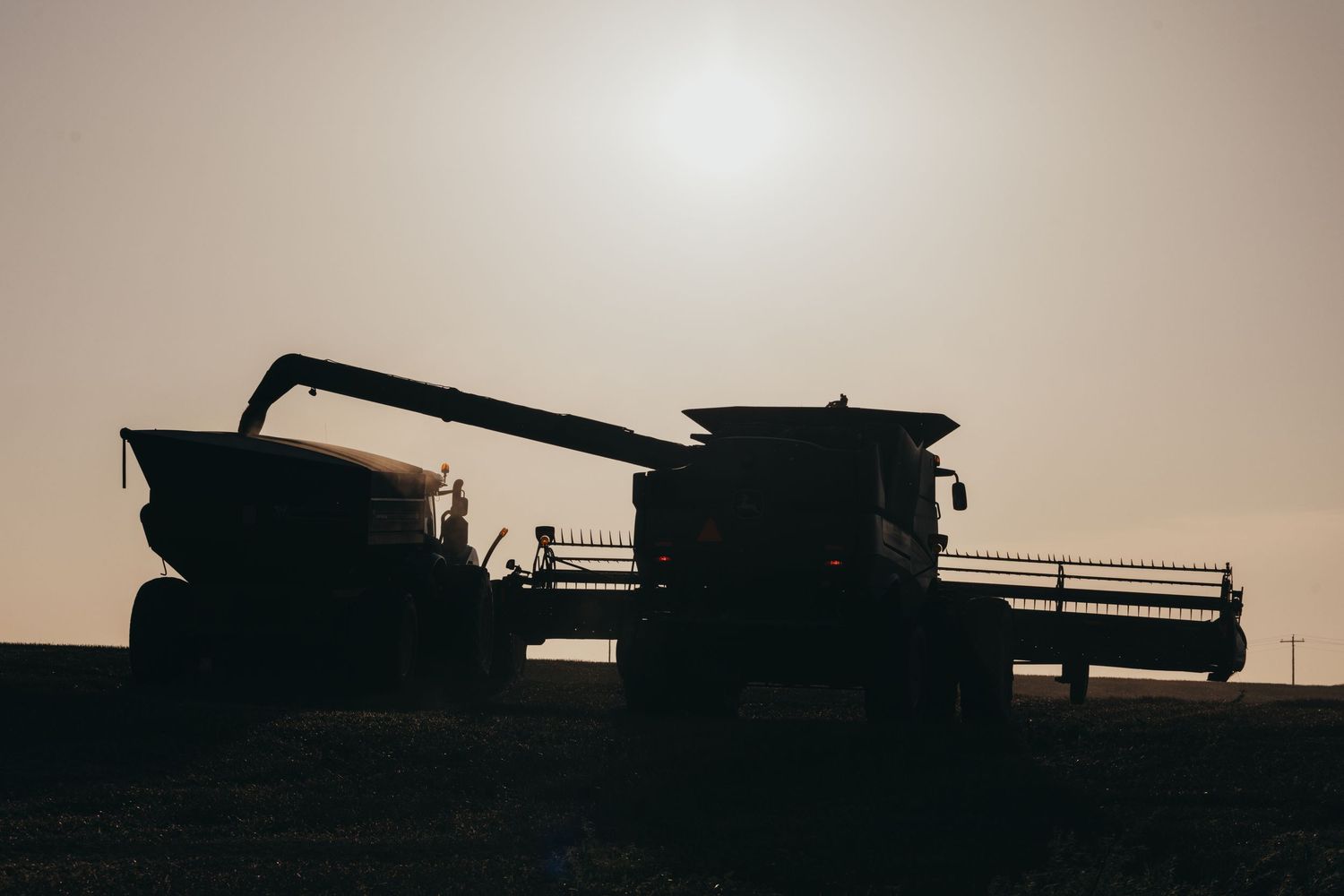 Silhouettes of farm machinery and combines working in a harvesting field during sunset create a dramatic agricultural scene.