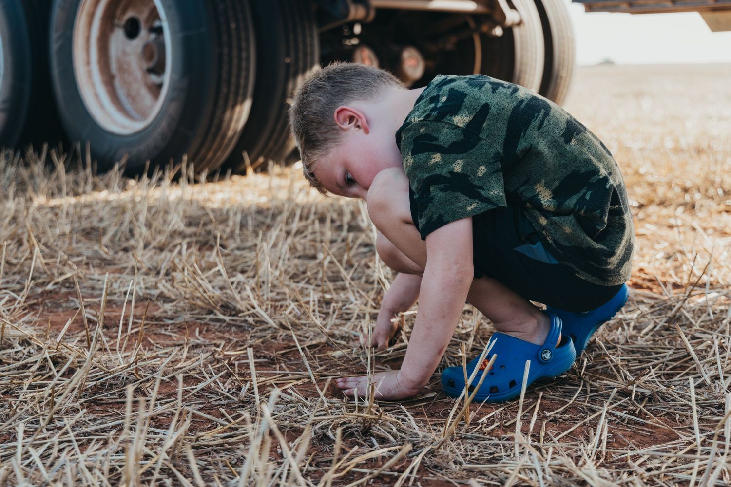 A small figure in a camo shirt and blue shorts crouches to explore dry straw beneath a truck tire outdoors.