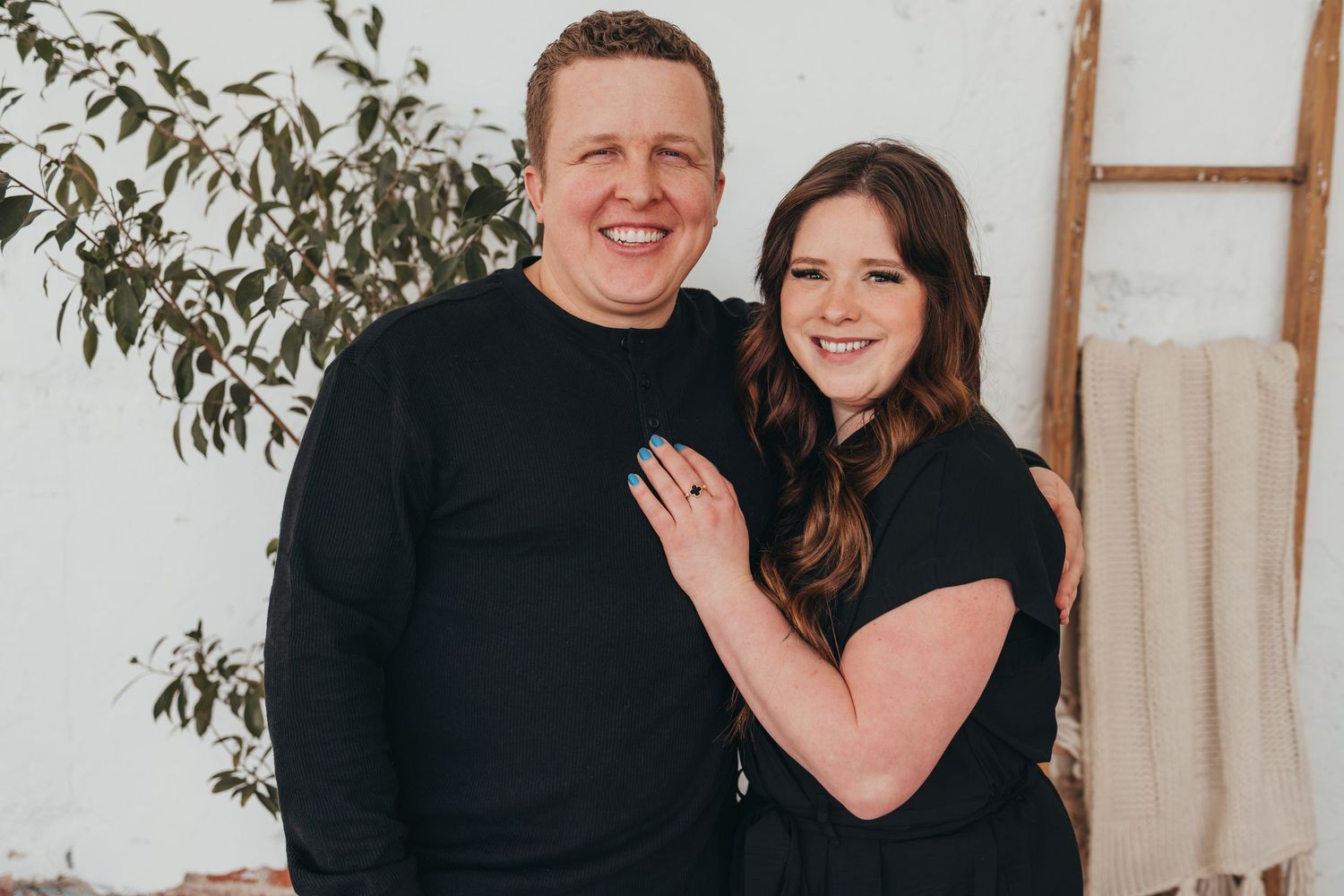 Couple in matching black outfits posing against white wall with greenery.