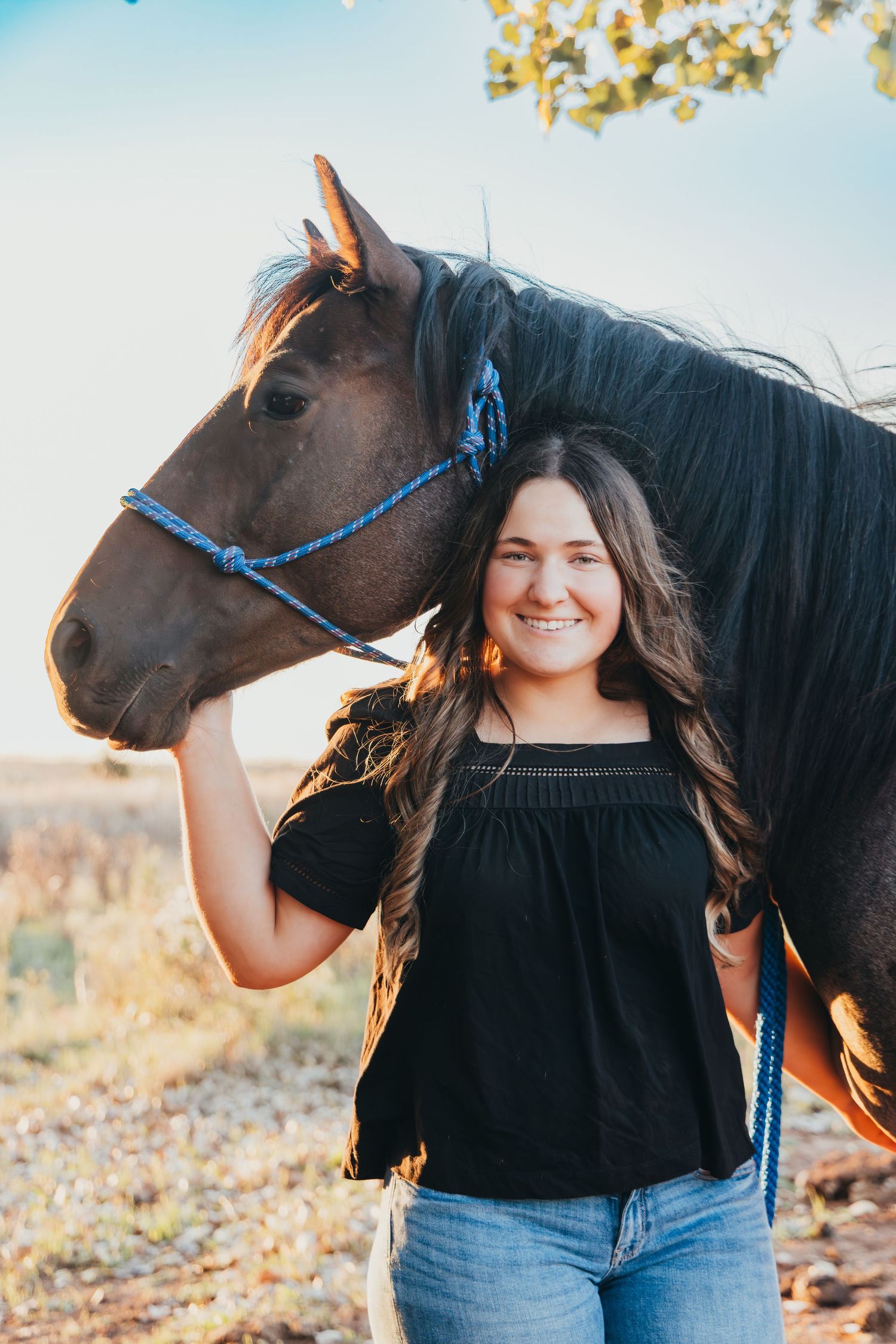 Smiling young woman in black top standing beside a dark brown horse with blue halter in a sunny field.