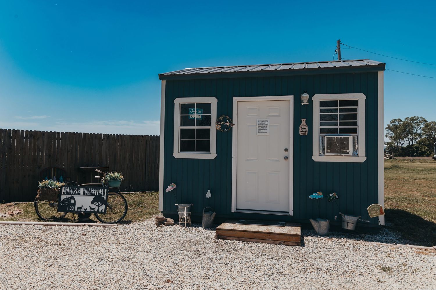 A small dark blue tiny house with white trim and potted plants sits on gravel with a bicycle parked outside.