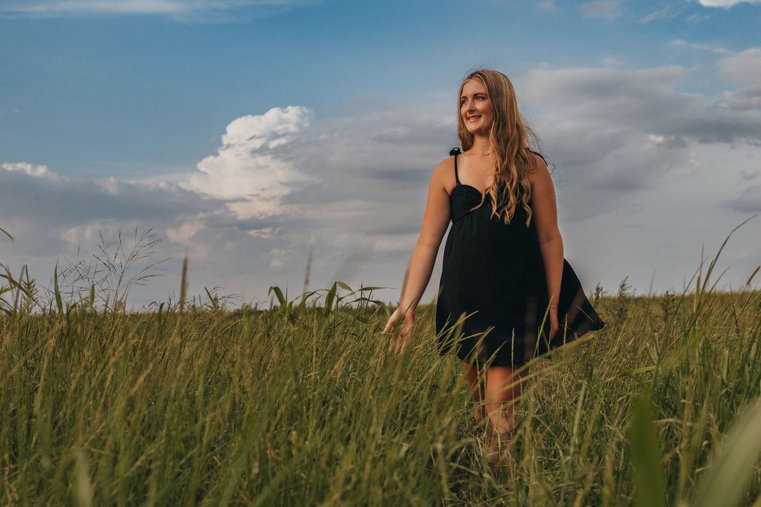 A figure stands among tall green wheat stalks in a rural field against a bright blue and cloudy summer sky.