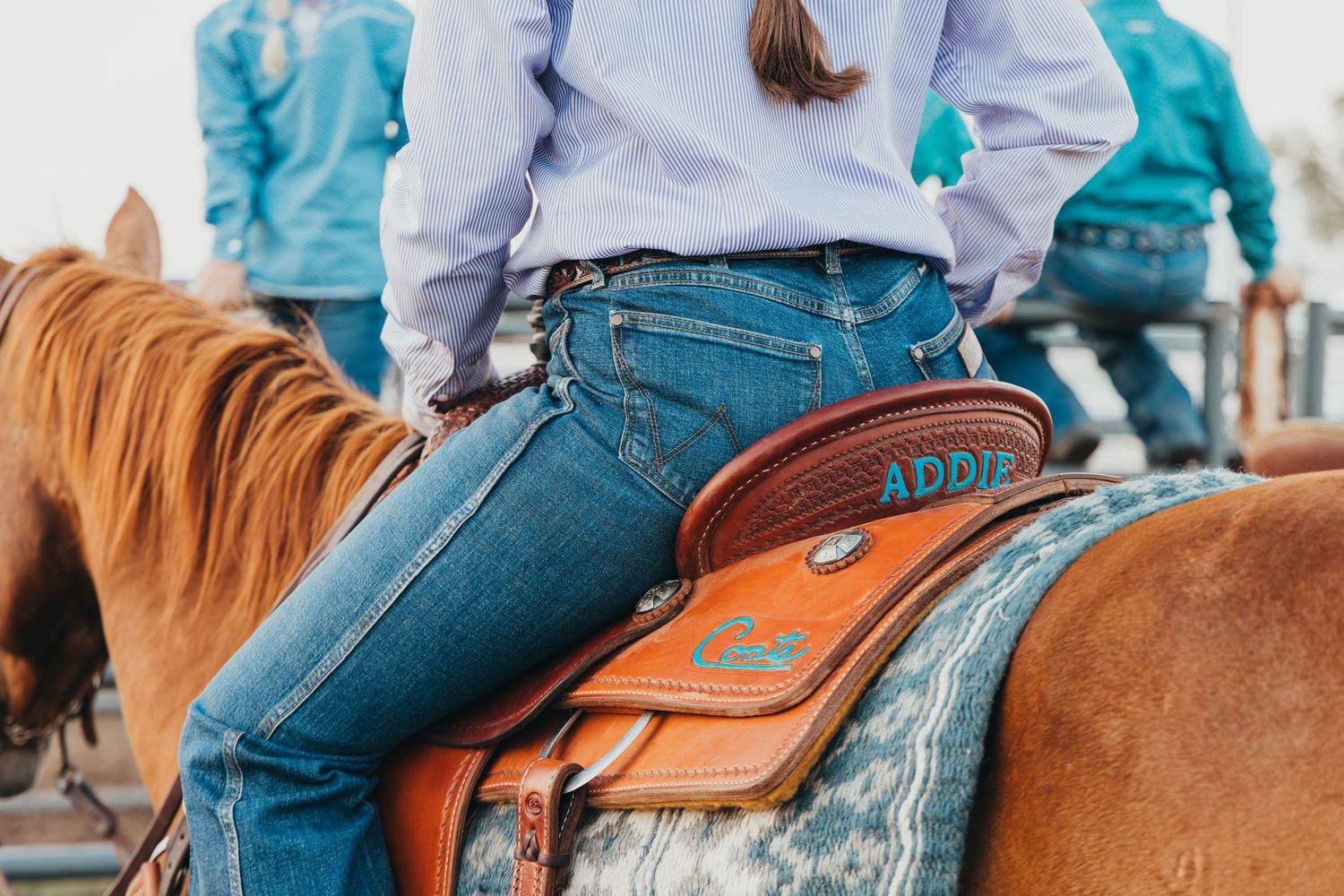 Close-up view of a rider in blue jeans sitting on a western saddle atop a chestnut horse.