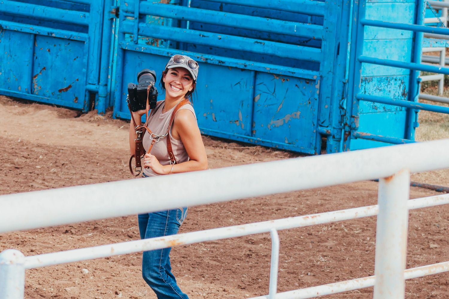 A photographer in jeans and a tank top smiles while leaning on a white railing near bright blue rodeo chutes.