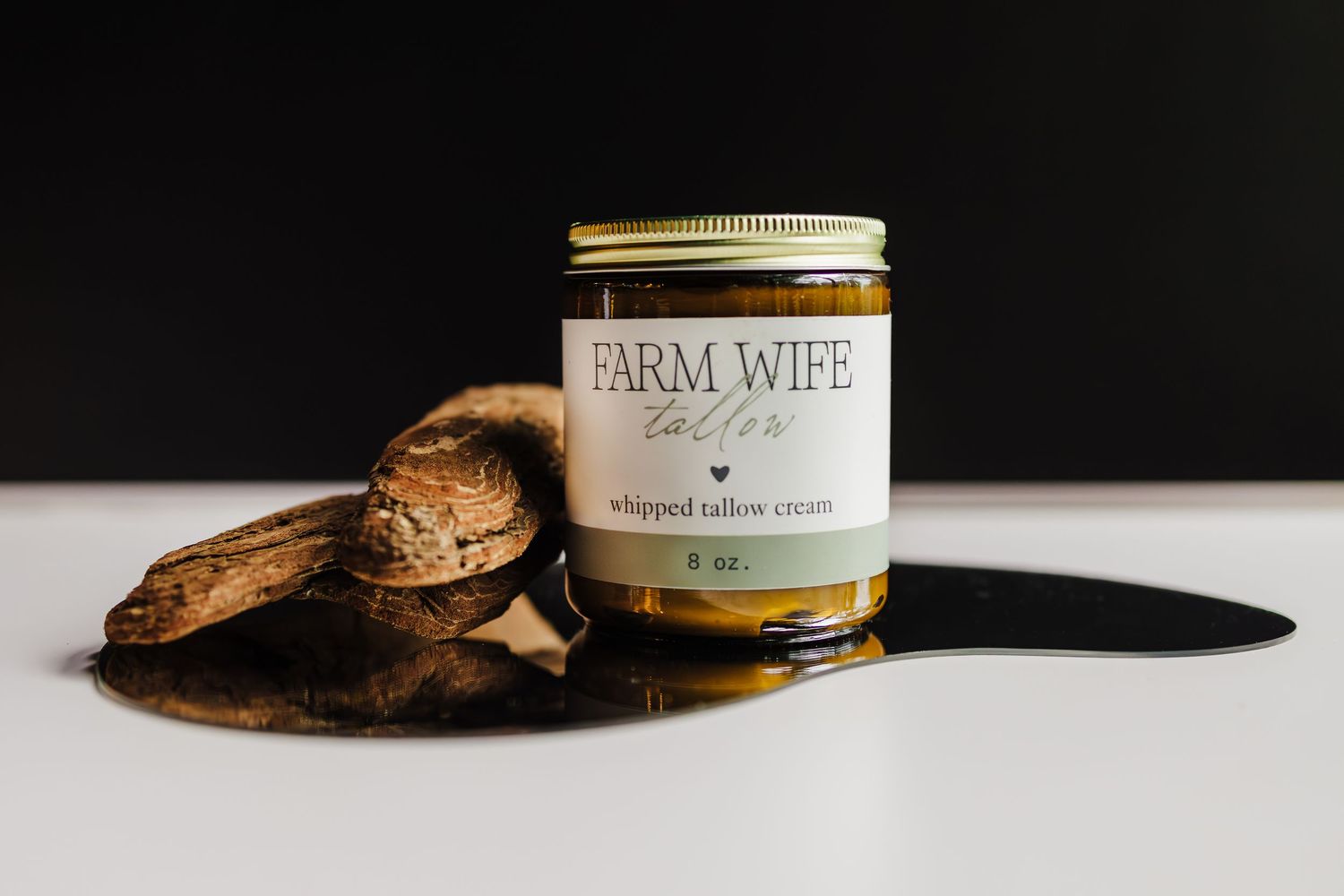A Farm Wife brand candle jar sits beside rustic wood on a white surface with dramatic shadow.