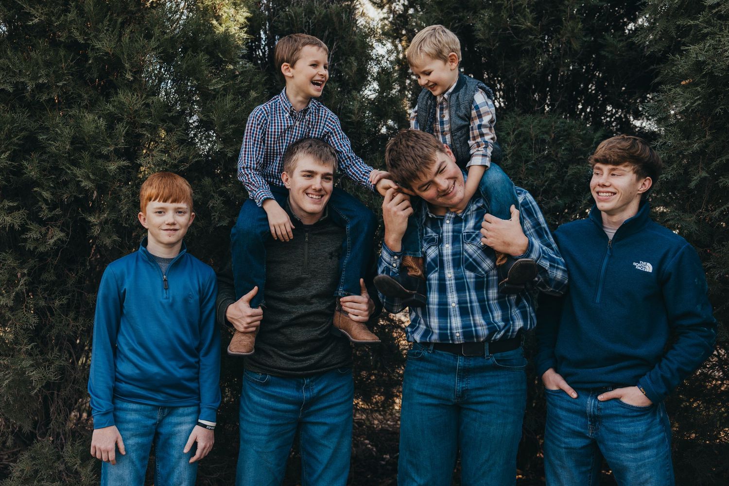 Group of young people in casual wear pose playfully together against evergreen trees.